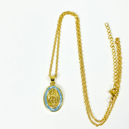 Gold chain necklace with an oval pendant featuring a gold religious medallion over a light blue patterned background, displayed on a plain white surface.