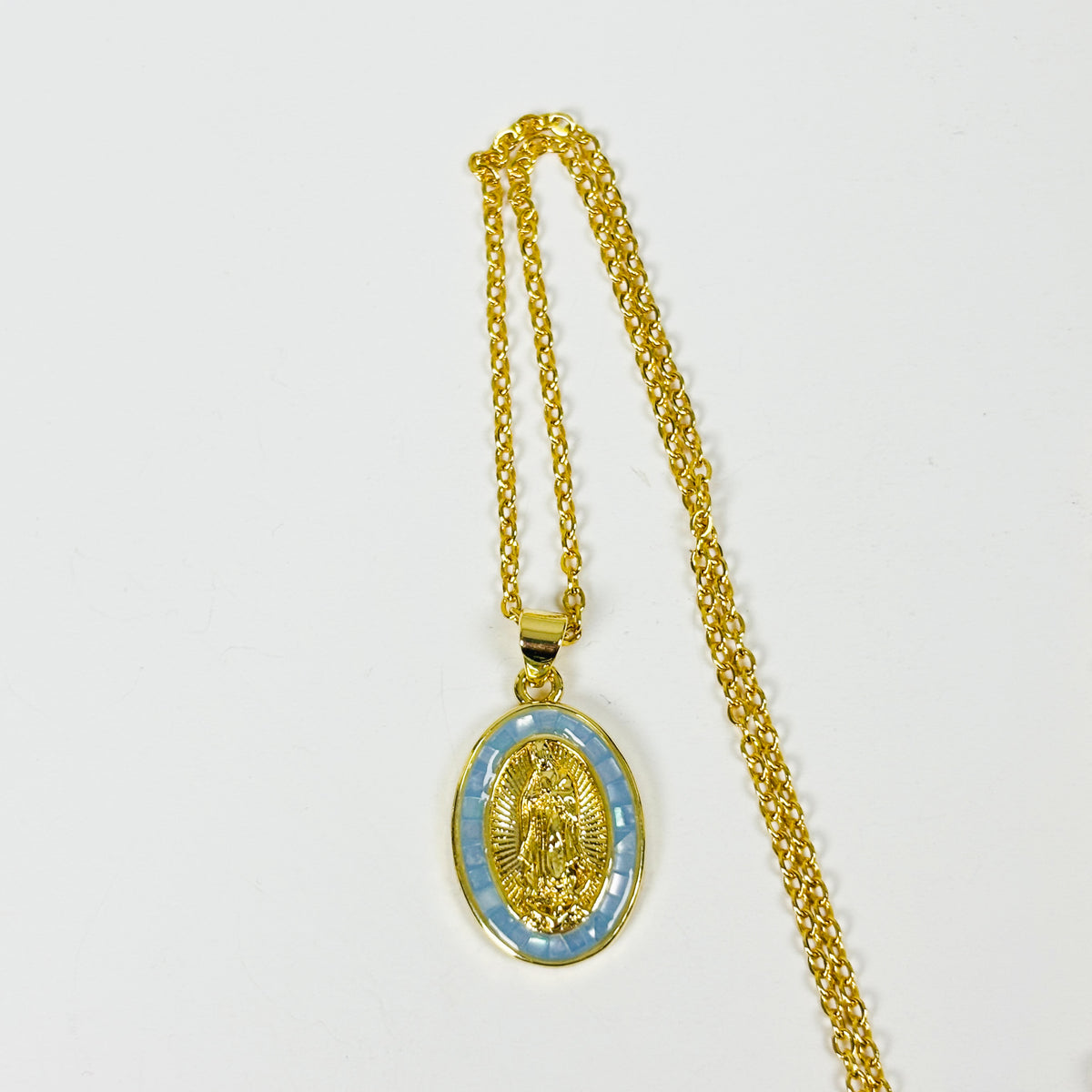 Gold chain necklace with an oval pendant featuring a gold religious medallion over a light blue patterned background, displayed on a plain white surface.