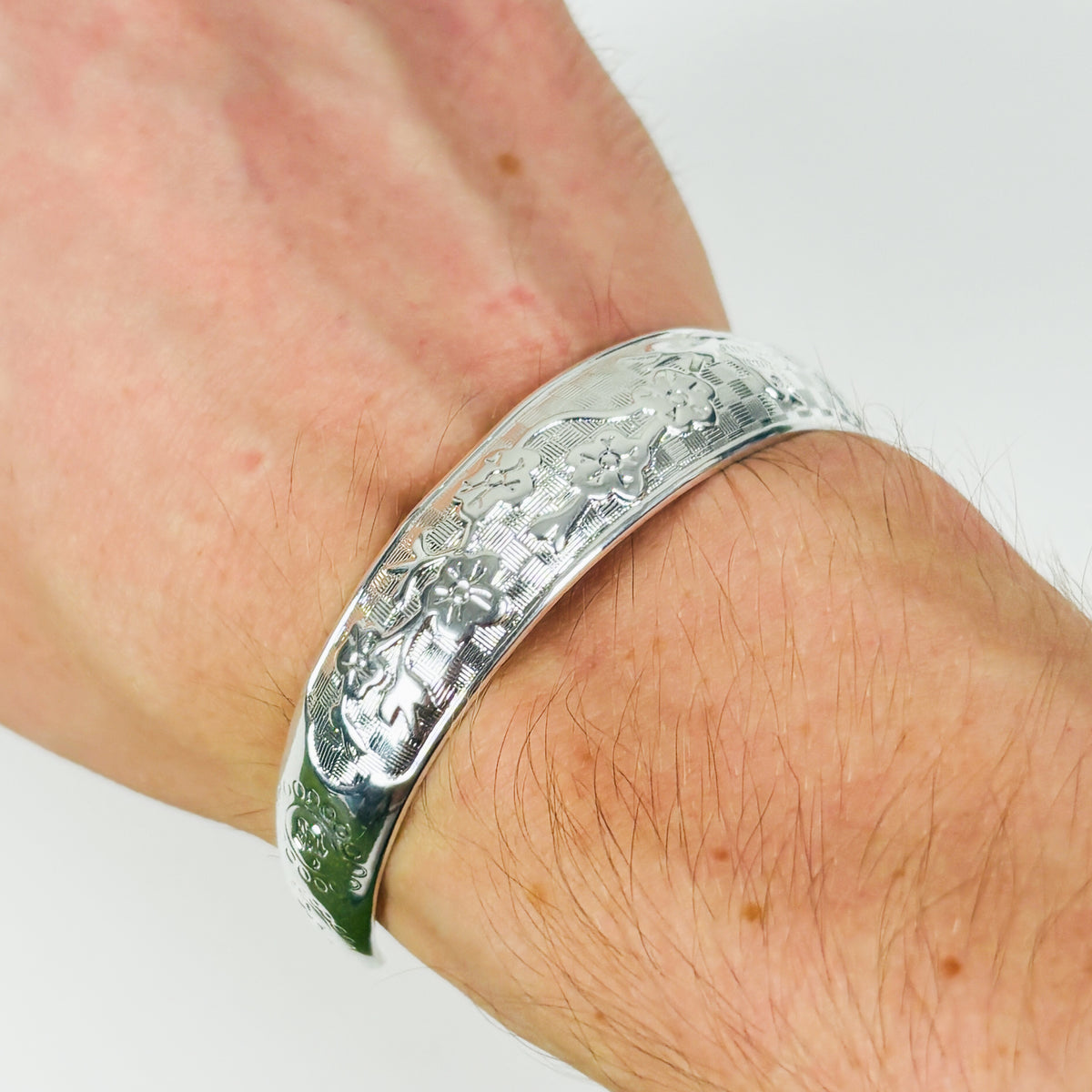 A close-up of a hand wearing a wide, polished silver bangle bracelet with an engraved floral or leaf pattern.